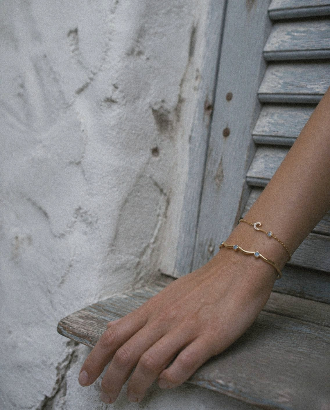 A hand resting on a pale blue shutter, wearing the Marea Bangle and the Onda Bracelet